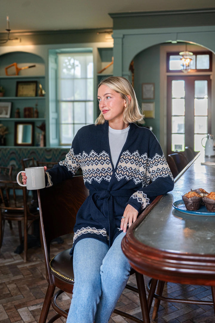 Woman in a cozy setting wearing a patterned sweater, sitting at a table with a cup and muffins.