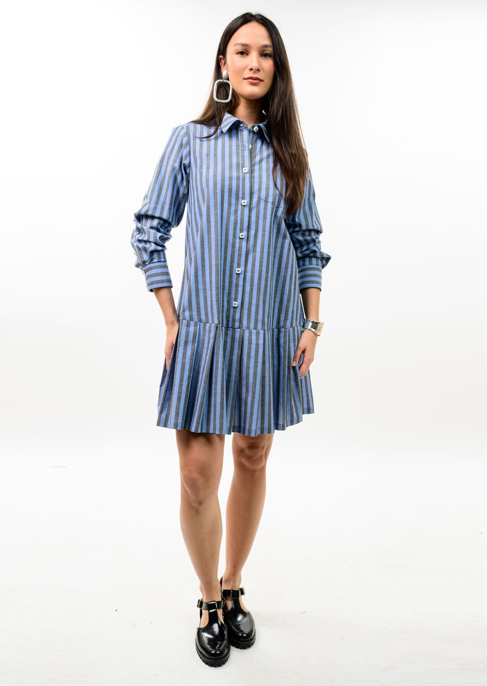 A woman modeling a blue and white vertically striped shirt dress with long sleeves, a pointed collar, and a tiered skirt, paired with black Mary Jane shoes and large silver hoop earrings, against a plain white studio background.