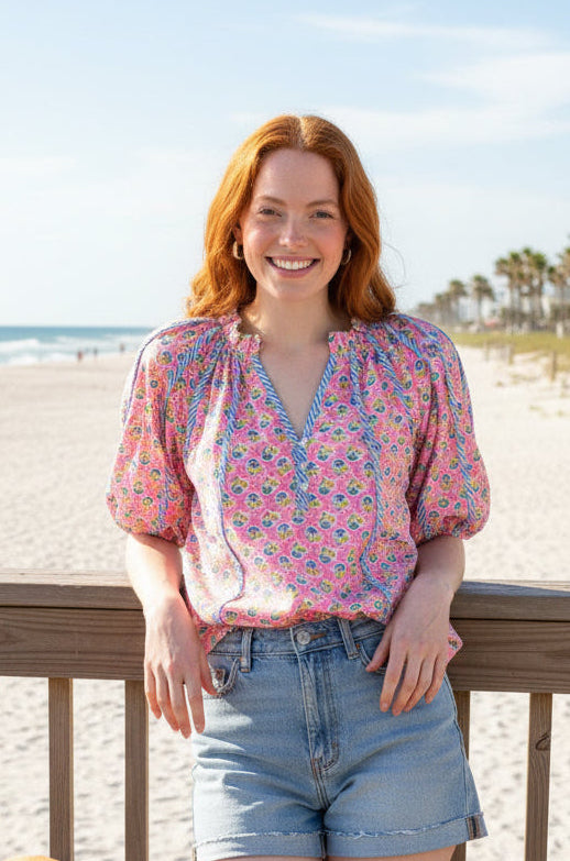 Woman in floral blouse and denim shorts standing on a beach with a dog by her side.