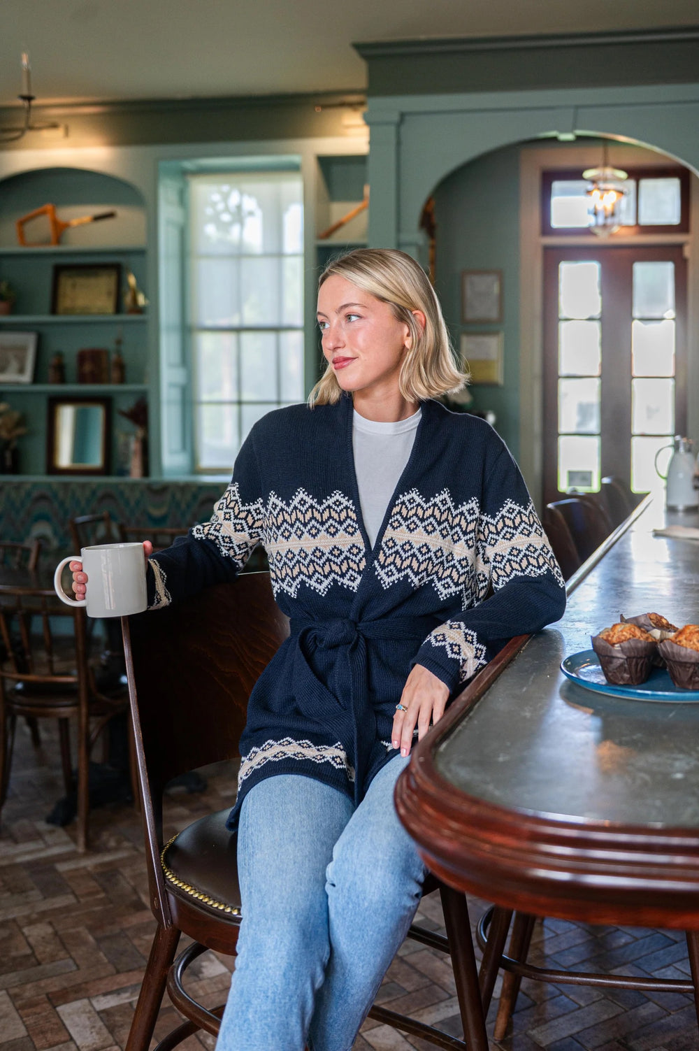 Woman in a cozy setting wearing a patterned sweater, sitting at a table with a cup and muffins.