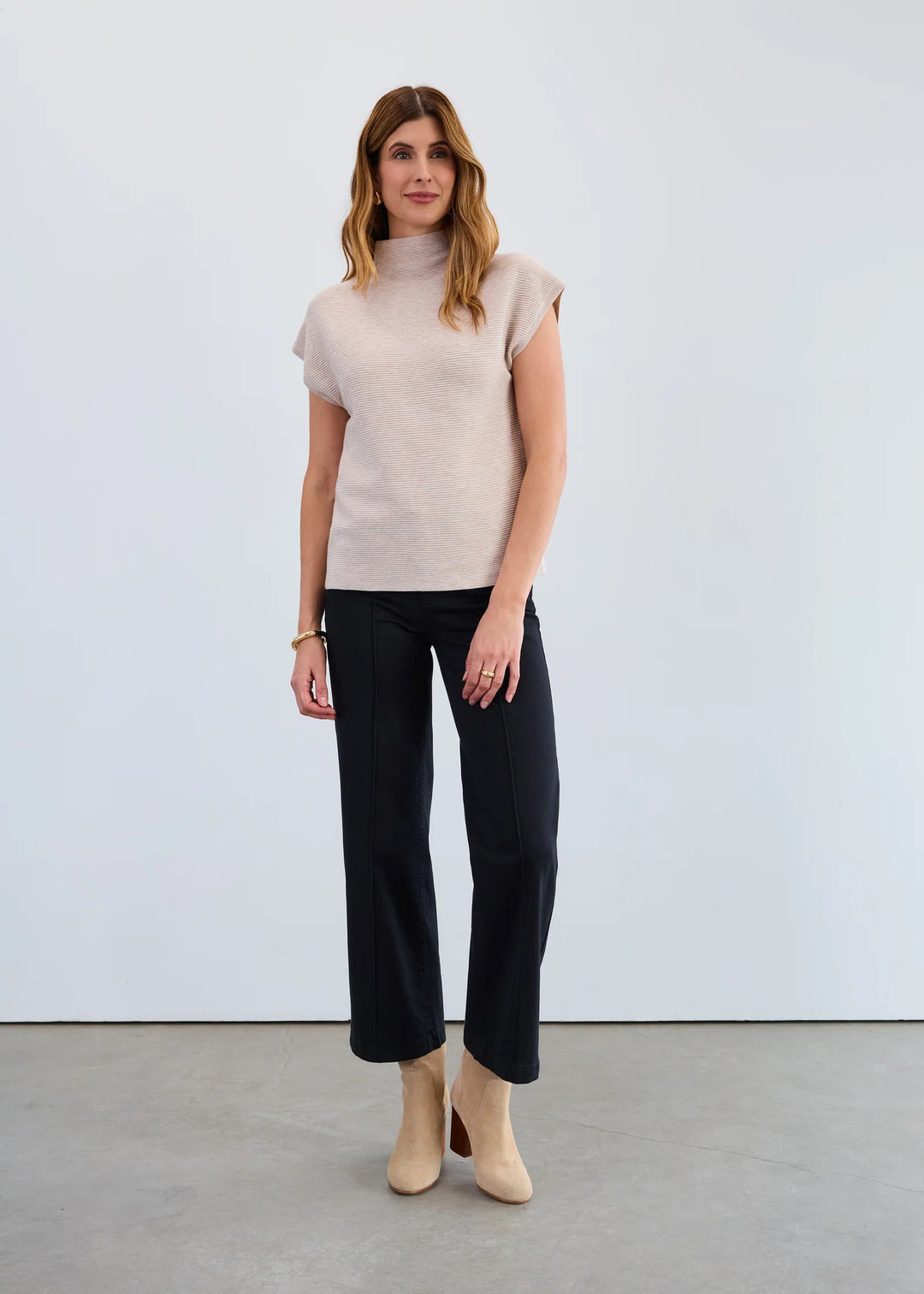 Woman wearing a beige turtleneck sweater and black pants against a white background