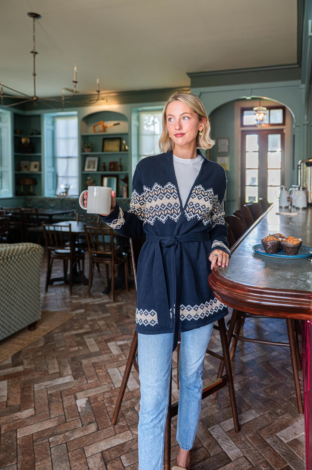 Woman in a cozy room holding a mug, wearing a navy cardigan with white patterns.